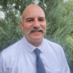 Matt Dawson (white, bald man with a mustache, wearing a light blue shirt and dark blue tie) smiles at the camera outside in front of foliage