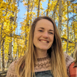 Mariama (woman with pale skin and brown hair, wearing a pink sweater and denim overalls) smiles at the camera in front of bright yellow aspens