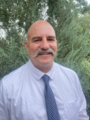 Matt Dawson (white, bald man with a mustache, wearing a light blue shirt and dark blue tie) smiles at the camera outside in front of foliage