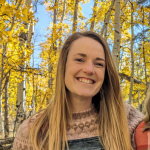 Mariama (woman with pale skin and brown hair, wearing a pink sweater and denim overalls) smiles at the camera in front of bright yellow aspens